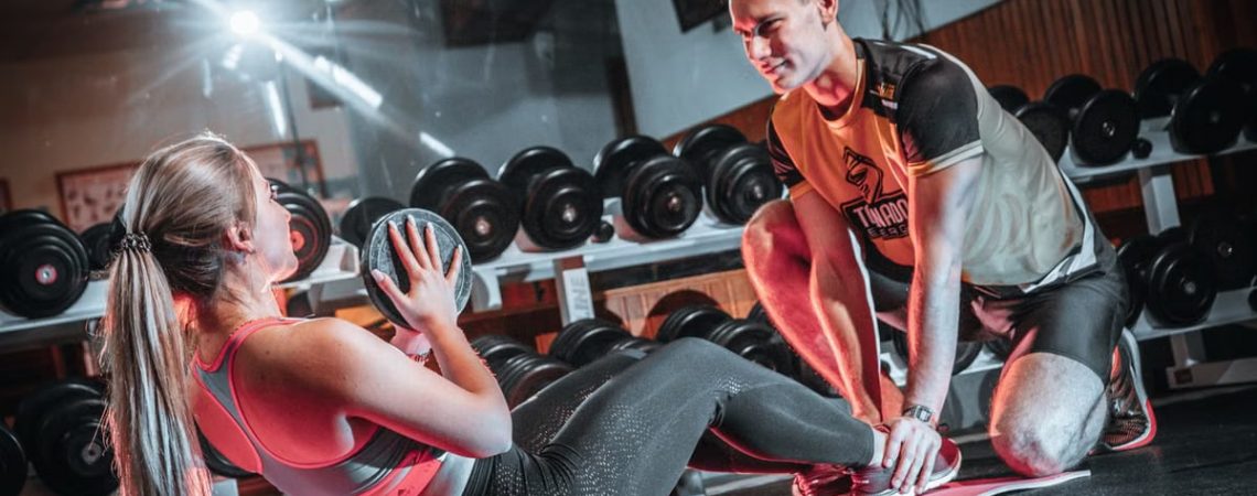 A personal trainer assists a woman doing sit-ups with a medicine ball in a gym setting.